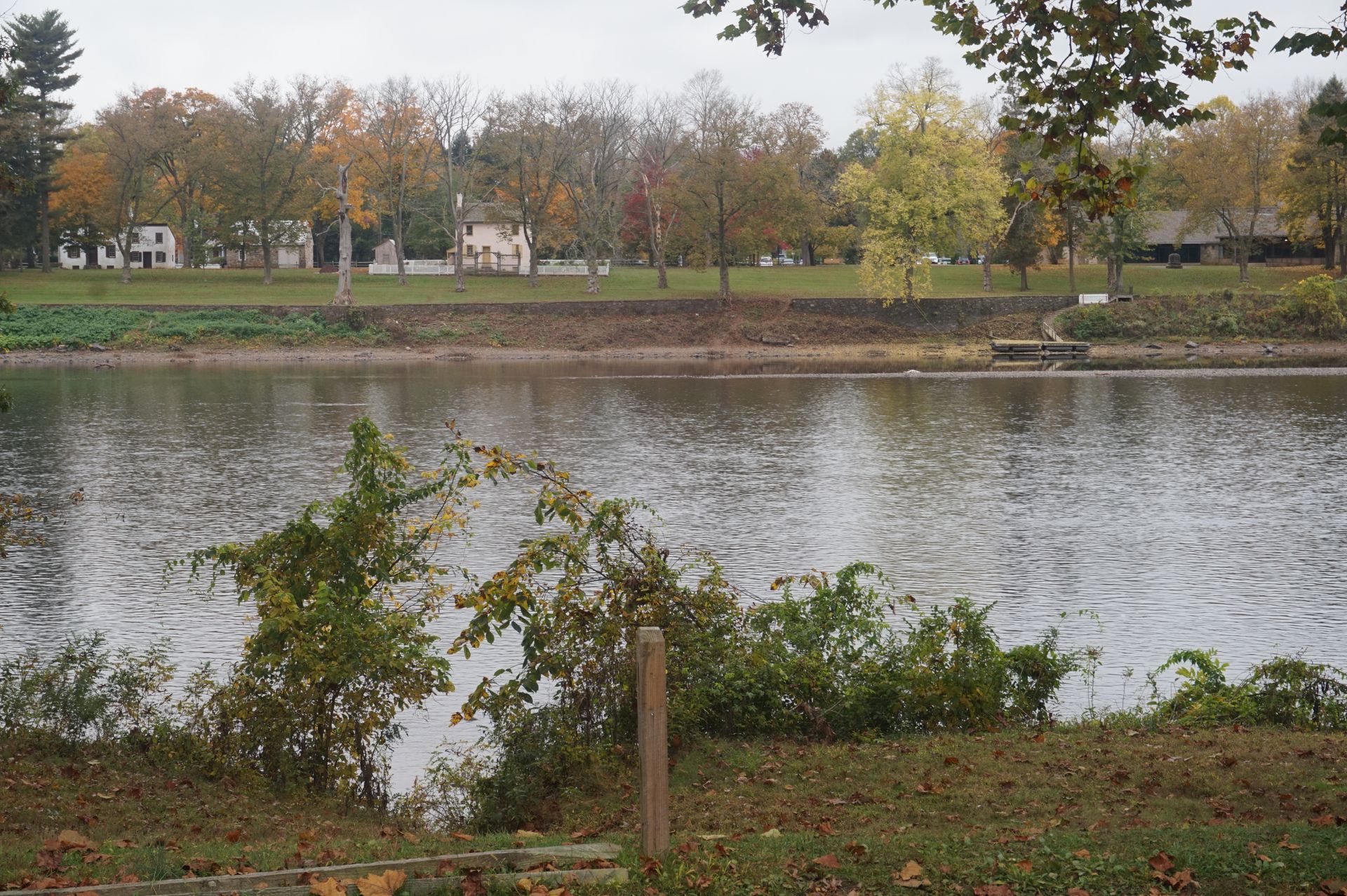A calm lake reflects trees with autumn colors. Houses line the far bank under an overcast sky.