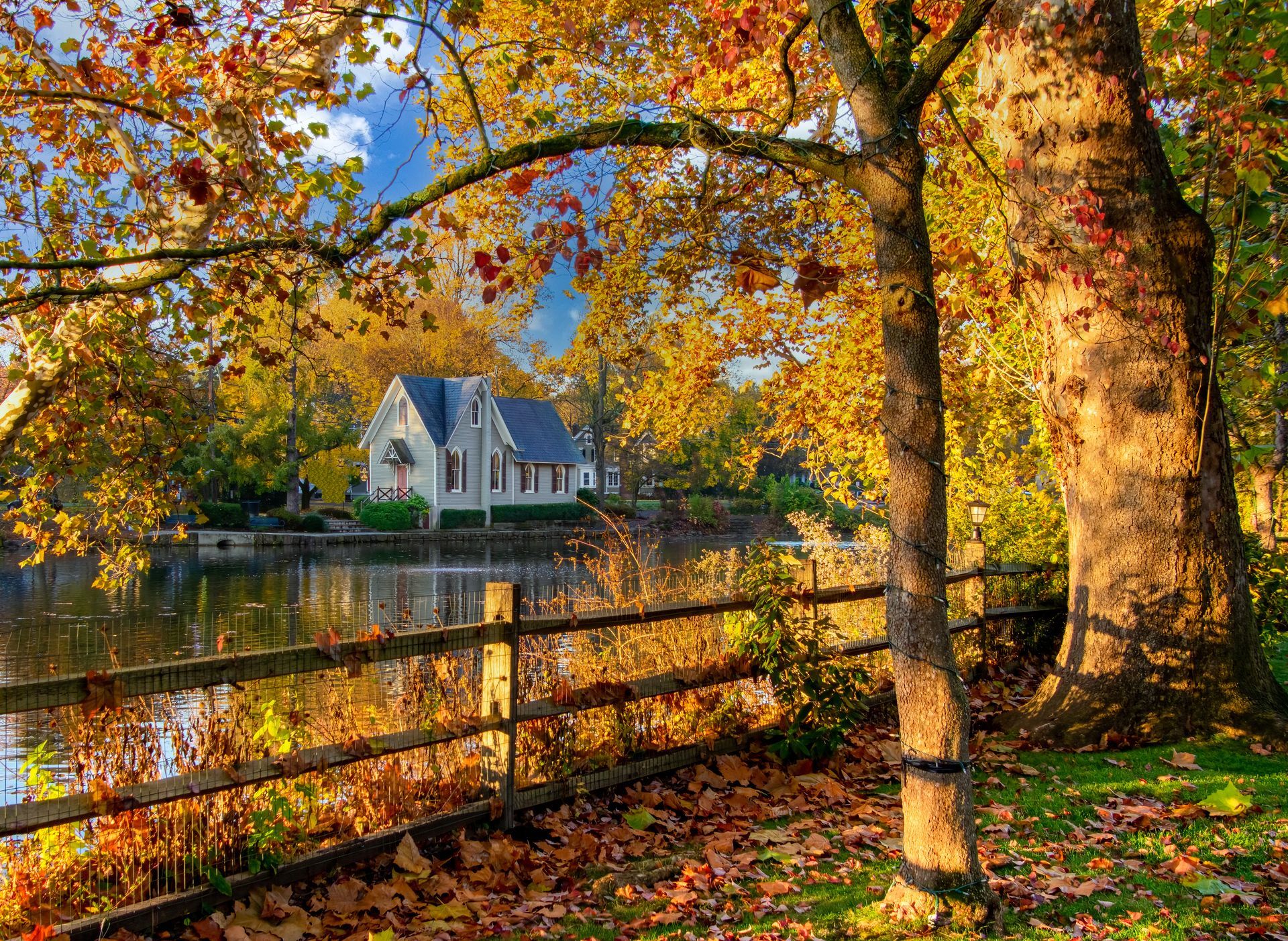 Autumn scene: a house by a lake with a wooden fence. Yellow leaves and trees frame the view under a blue sky.