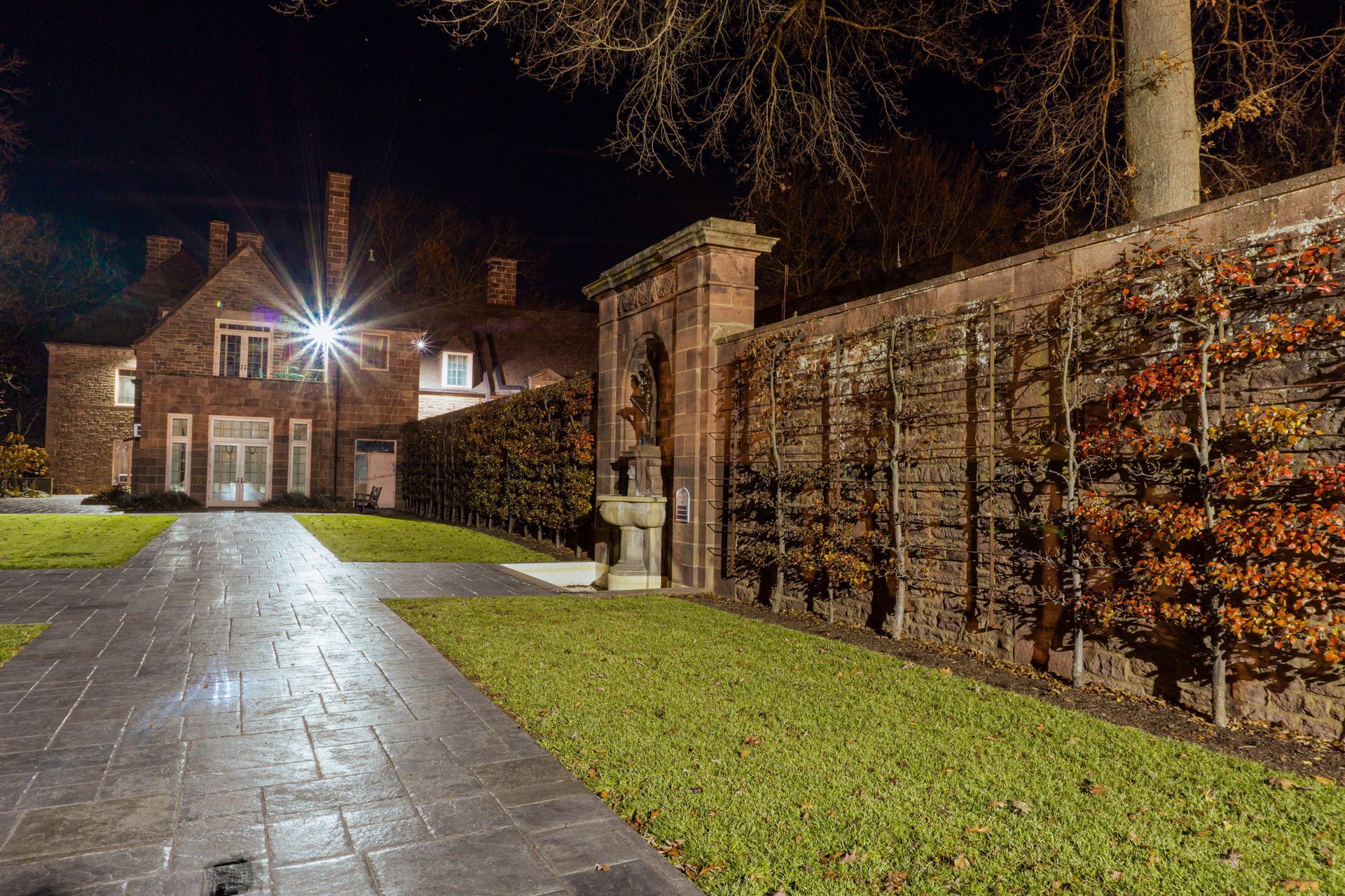 A brick house at night with a paved pathway, lit by a street lamp, and a hedge alongside the house.