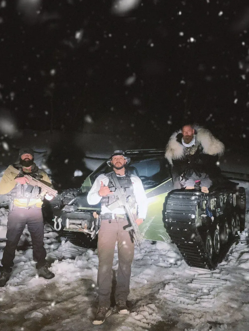 Three men in winter gear stand near a tracked vehicle in snow. Dark sky.