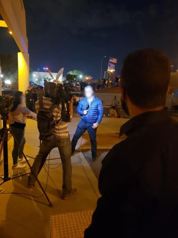 A reporter in a blue jacket speaks into a microphone outside at night, with a camera crew.