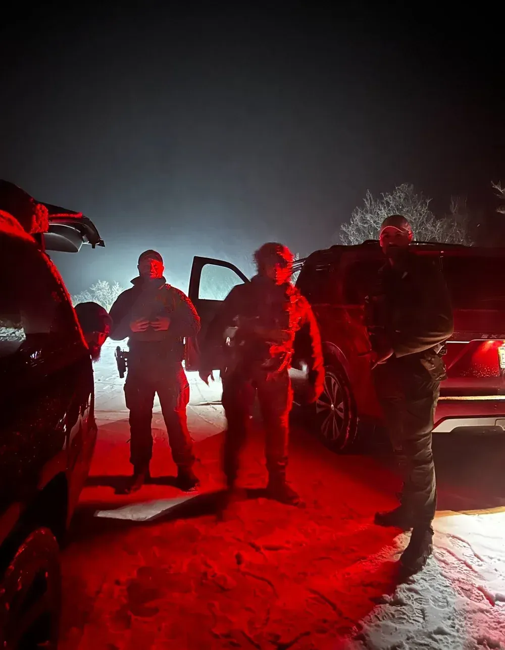 Men in tactical gear stand near vehicles at night, illuminated by red light. Snow is on the ground.