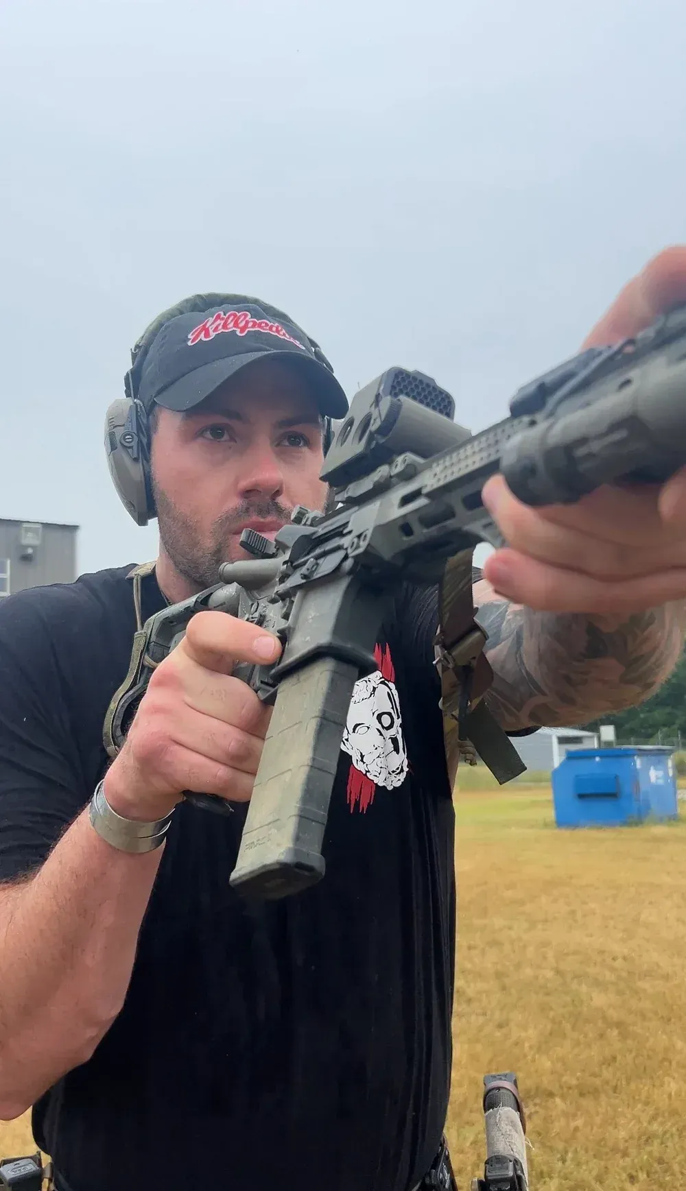 Man aiming a rifle at a range, wearing a hat, ear protection, and a black t-shirt.