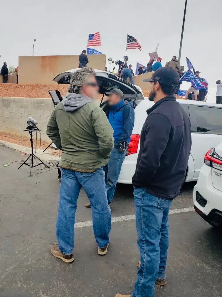 Three men in a parking lot with American flags in the background; one is holding a microphone.