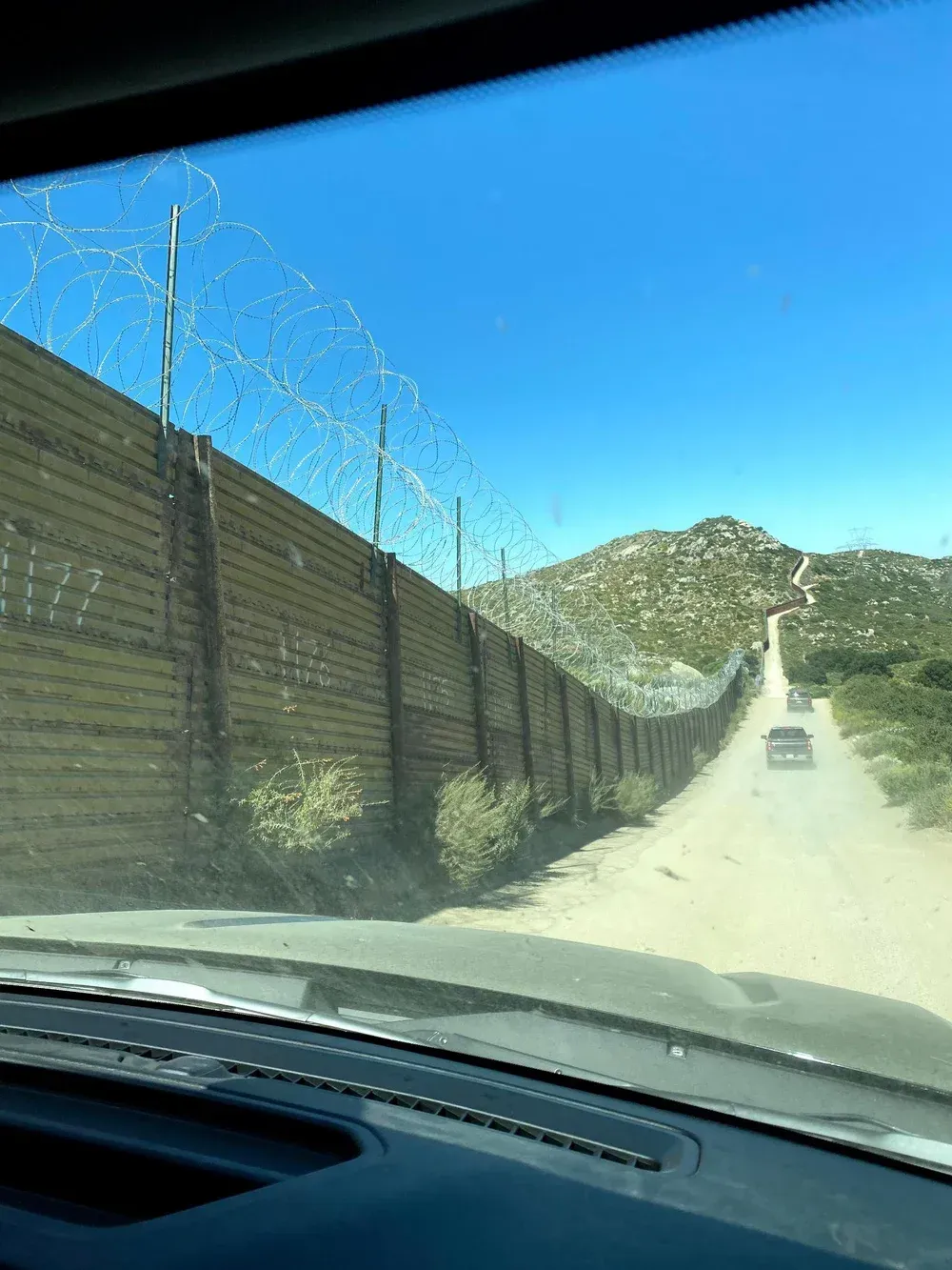 A vehicle driving down a dirt road along a border wall topped with razor wire under a blue sky.