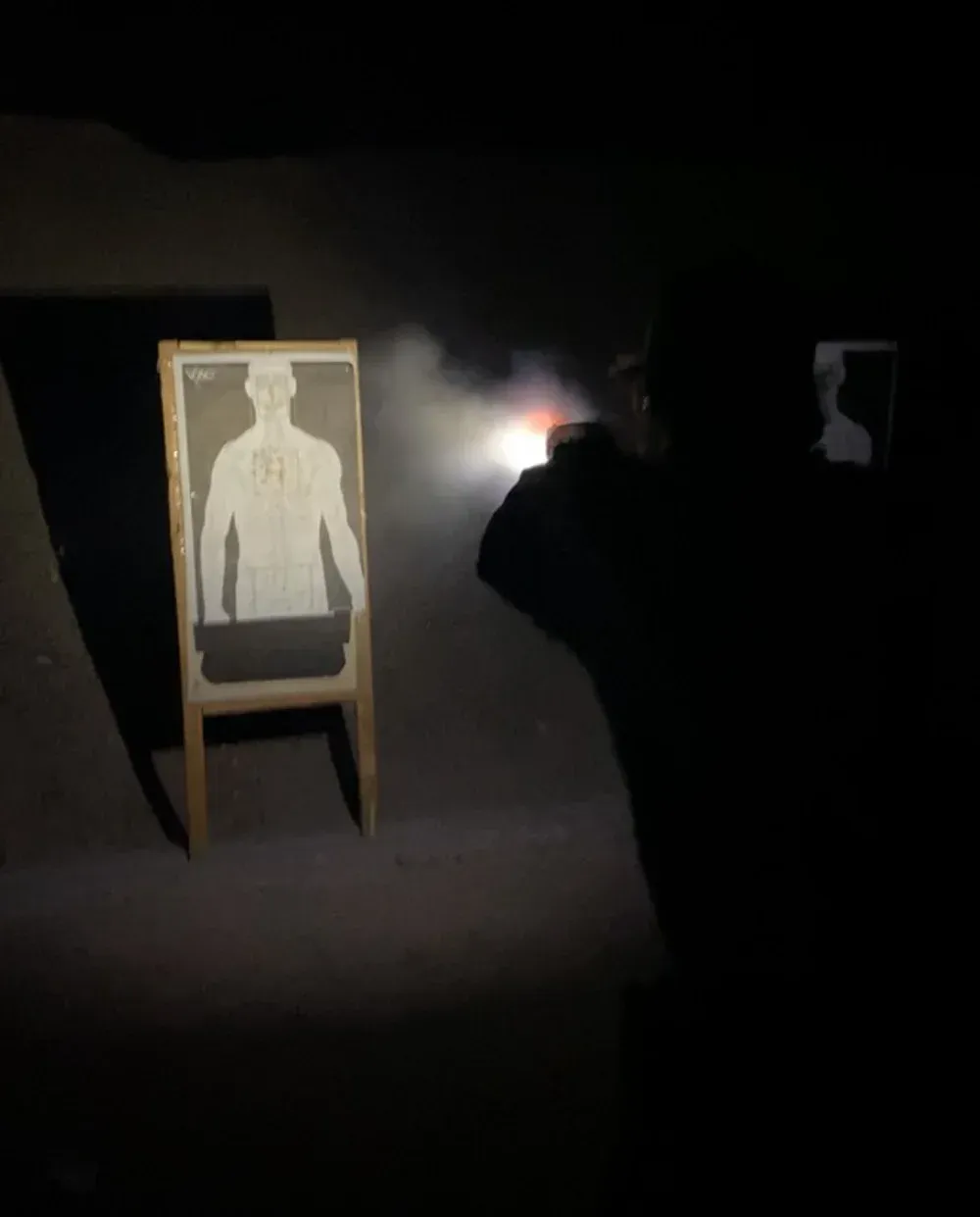 Person shooting a gun at a target in a dark indoor shooting range. Flash of the gun's blast illuminates the scene.