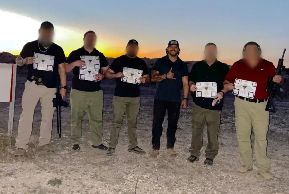 Six men at a firing range, holding targets. Sunset in the background. Some men have weapons.