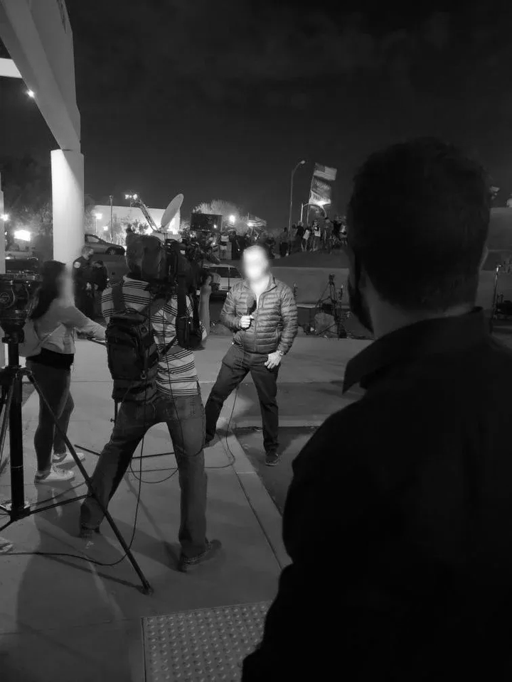 Journalist reporting outdoors at night, with a camera and crew nearby.