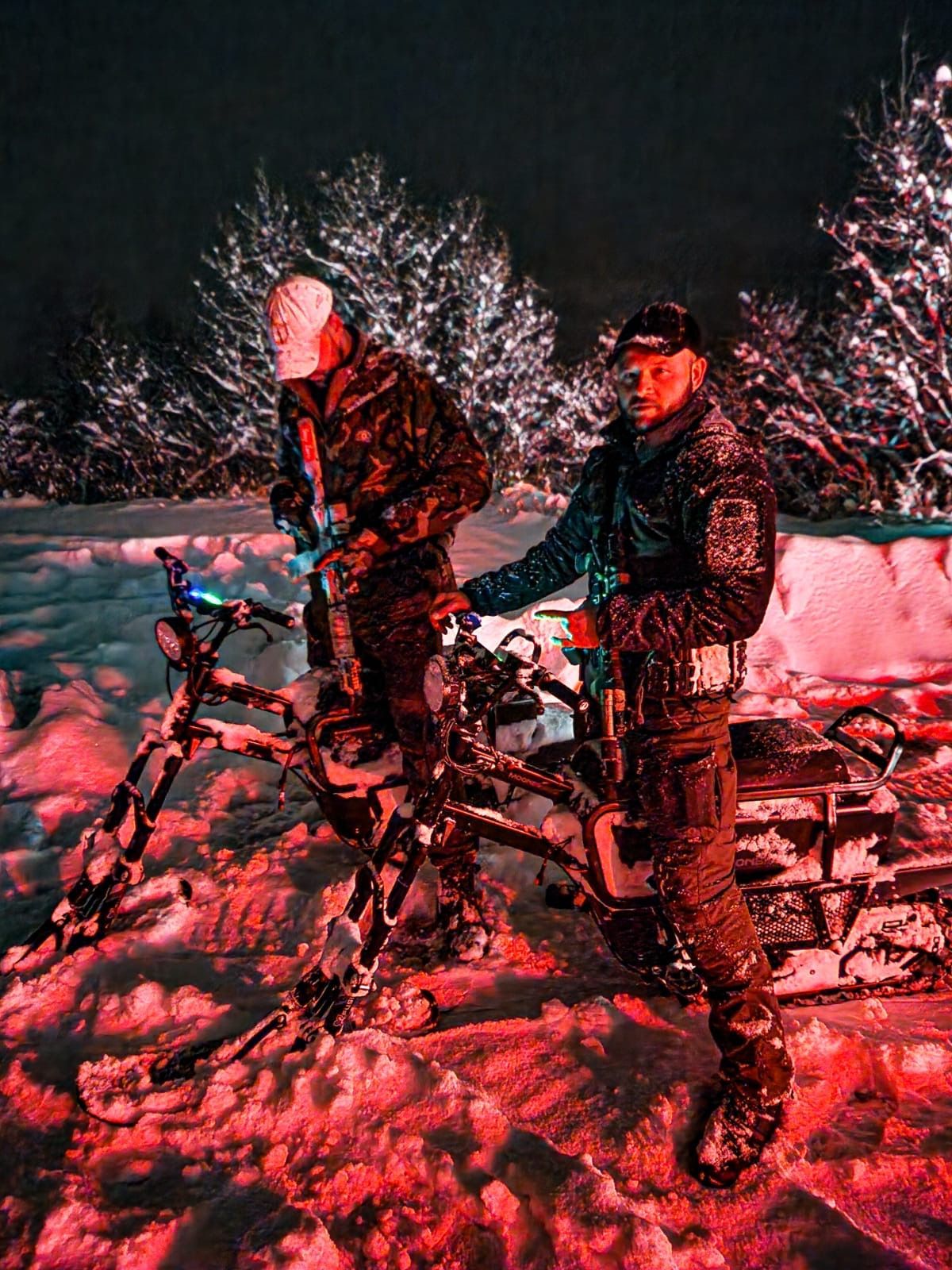 Two police officers stand in snow at night, illuminated by red light. One adjusts a snowmobile.