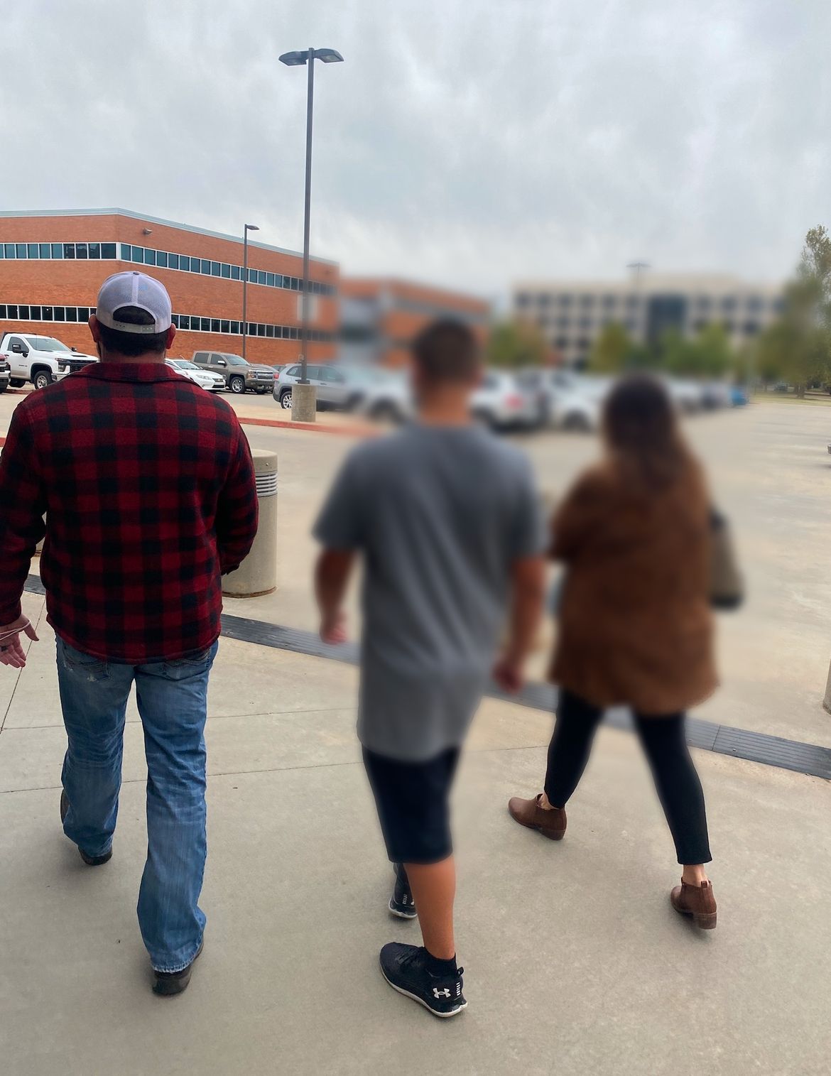 Three people walking away from the camera, parking lot in the background. Man in plaid shirt, others blurred.
