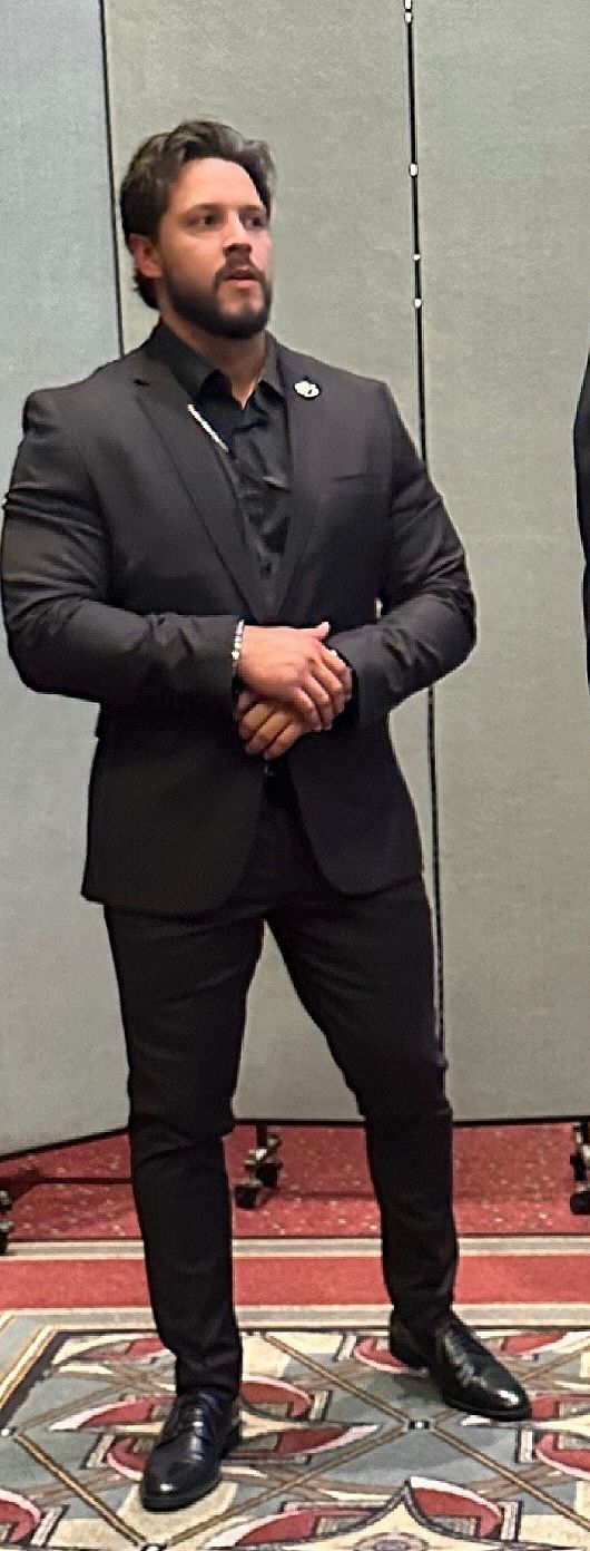 Man in a dark suit, standing with hands clasped in front of him, indoors.