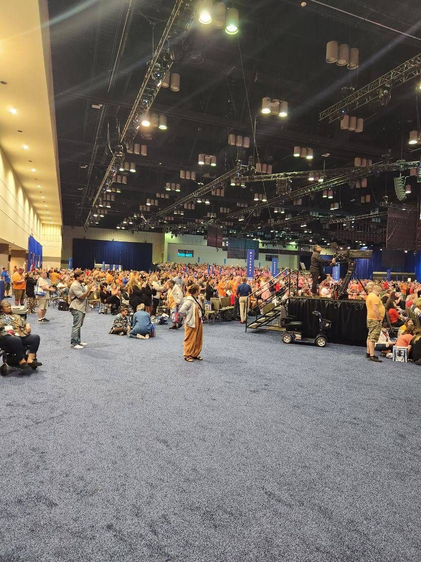 Large convention hall, crowd of people in orange shirts, blue carpet, stage in the background.