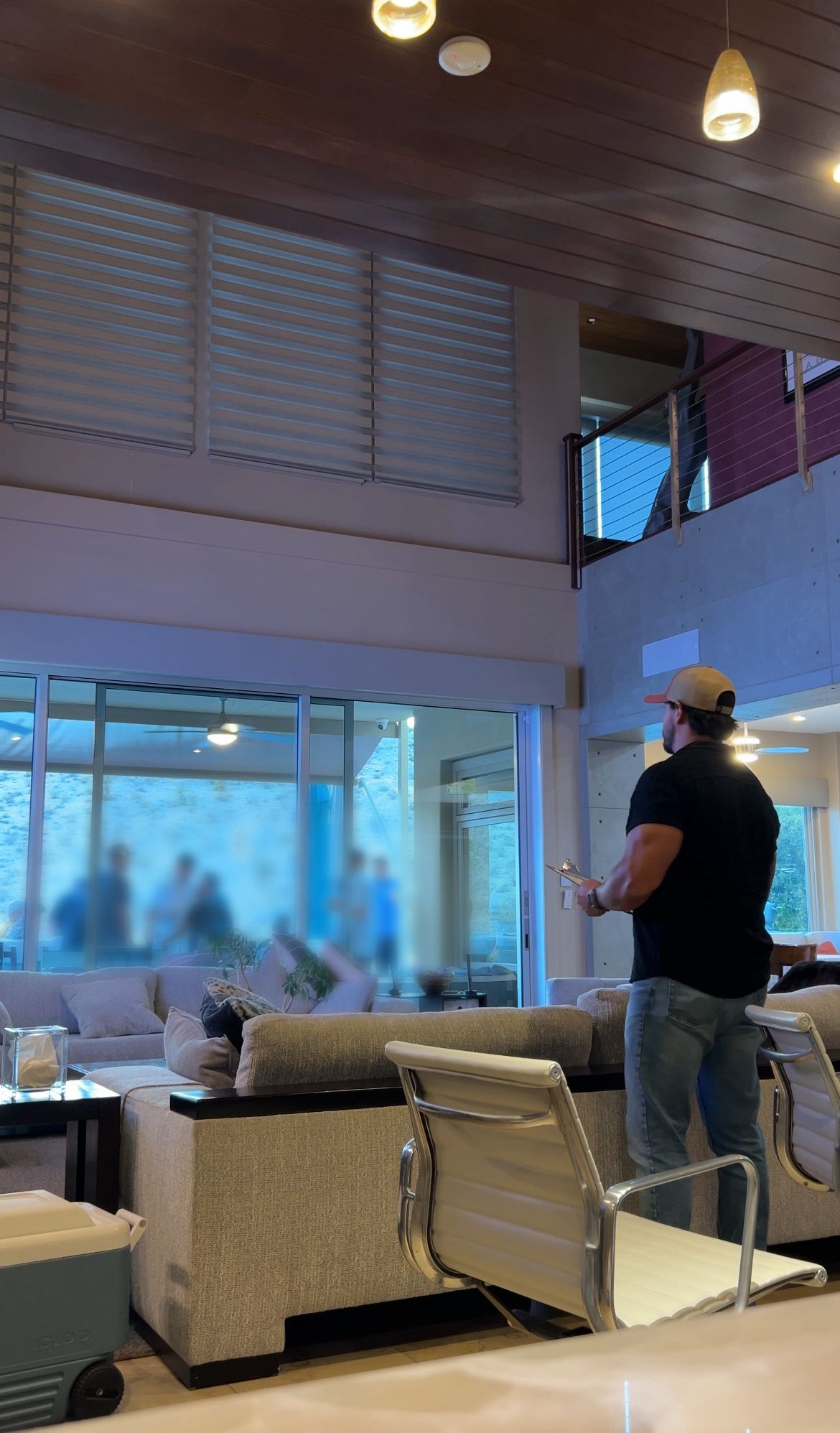 Man in a cap holding something indoors, standing near a window looking at a blurred group; modern interior.