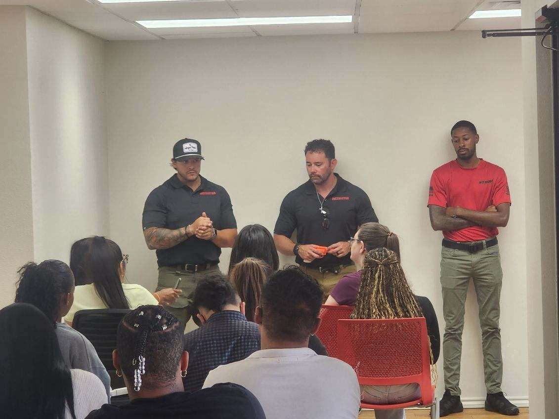 Men giving a presentation to a seated audience in a room; one man has a tattoo.