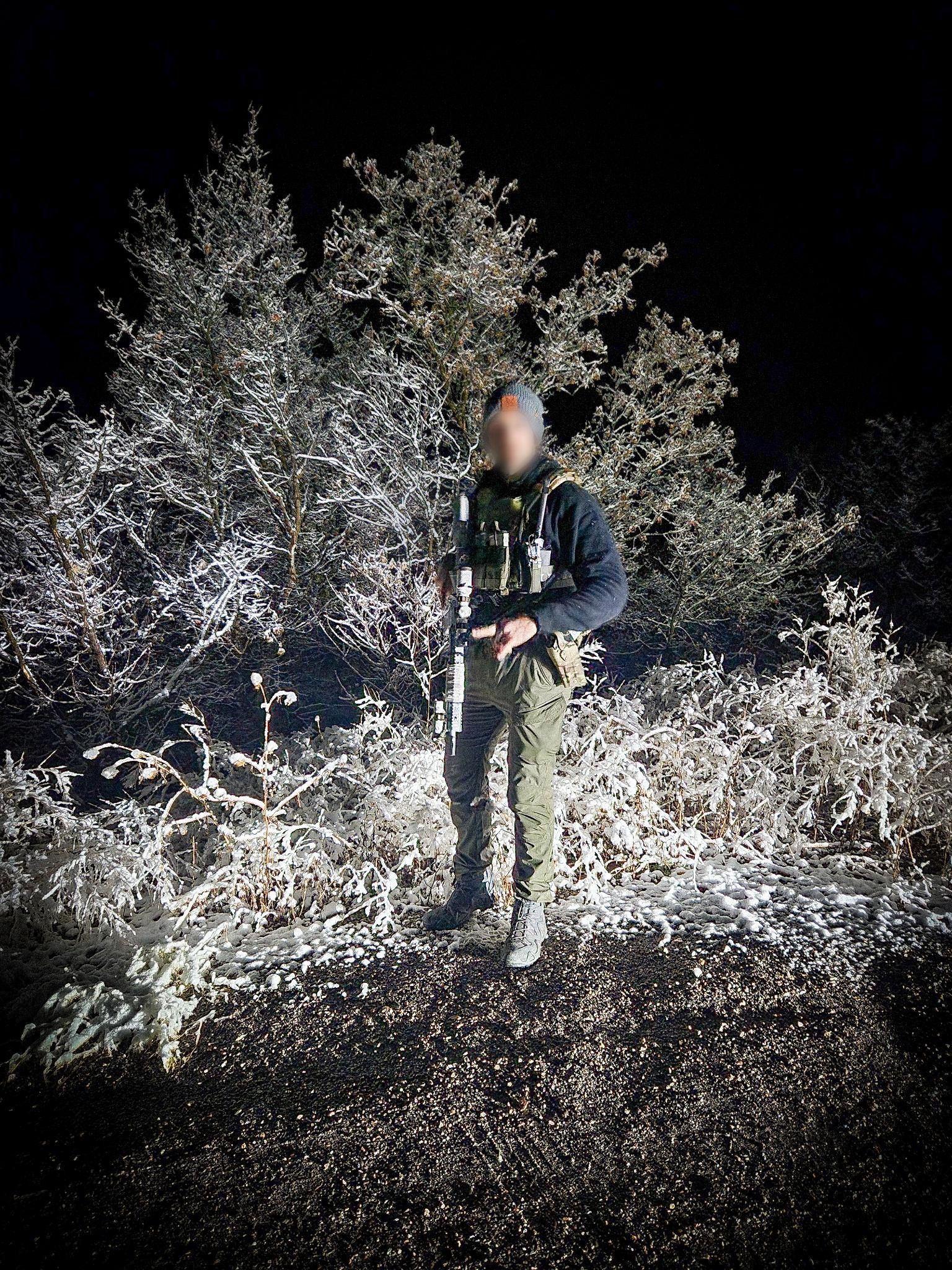 Person in camouflage gear stands outdoors at night, illuminated by a bright light.