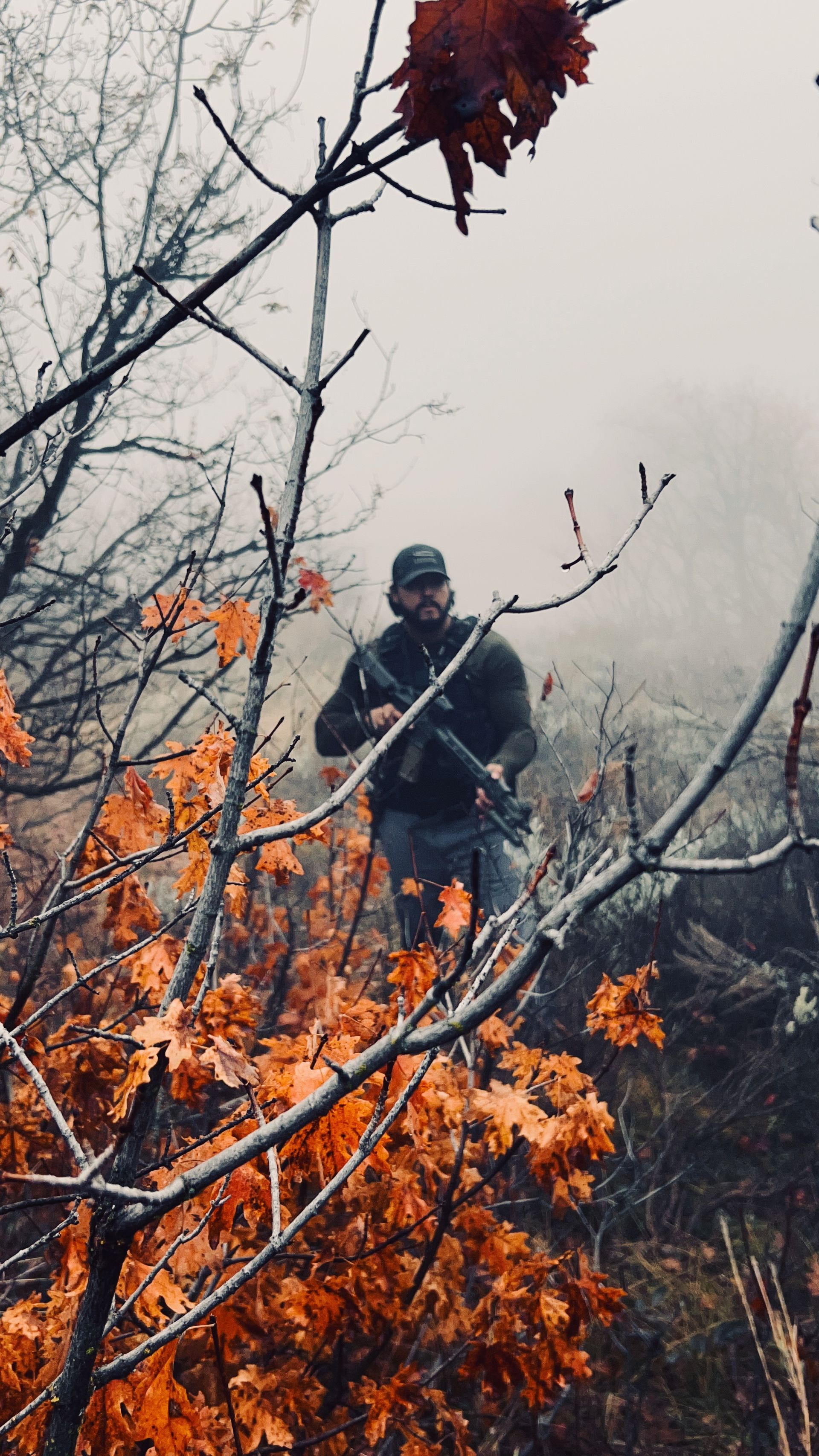 Man holding a rifle in a foggy forest with orange leaves.