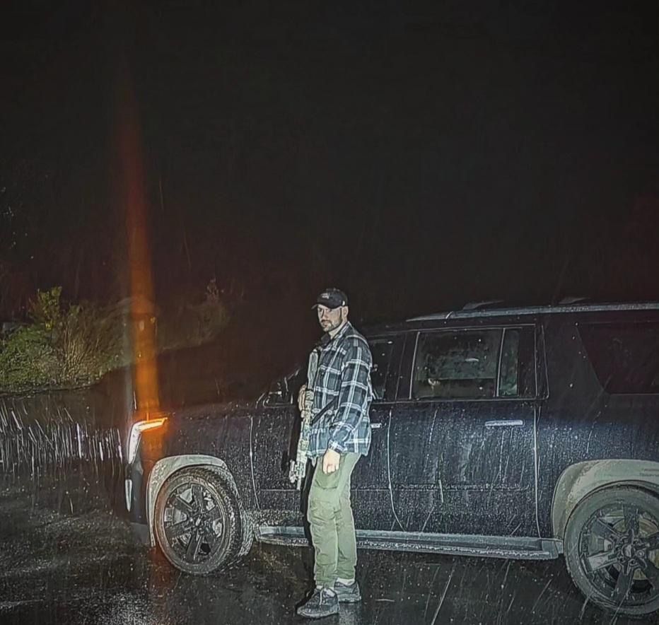 Man in plaid shirt and cap stands by black SUV at night, holding a rifle.