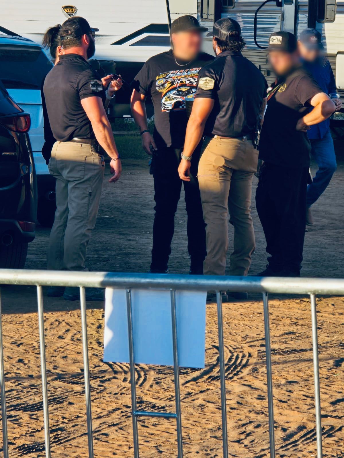 Group of men standing near a fence. Some are wearing black shirts, tan pants, and hats; others have blurred faces.