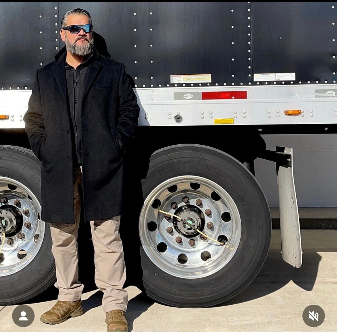 Man in coat and sunglasses standing next to the wheels of a large truck.