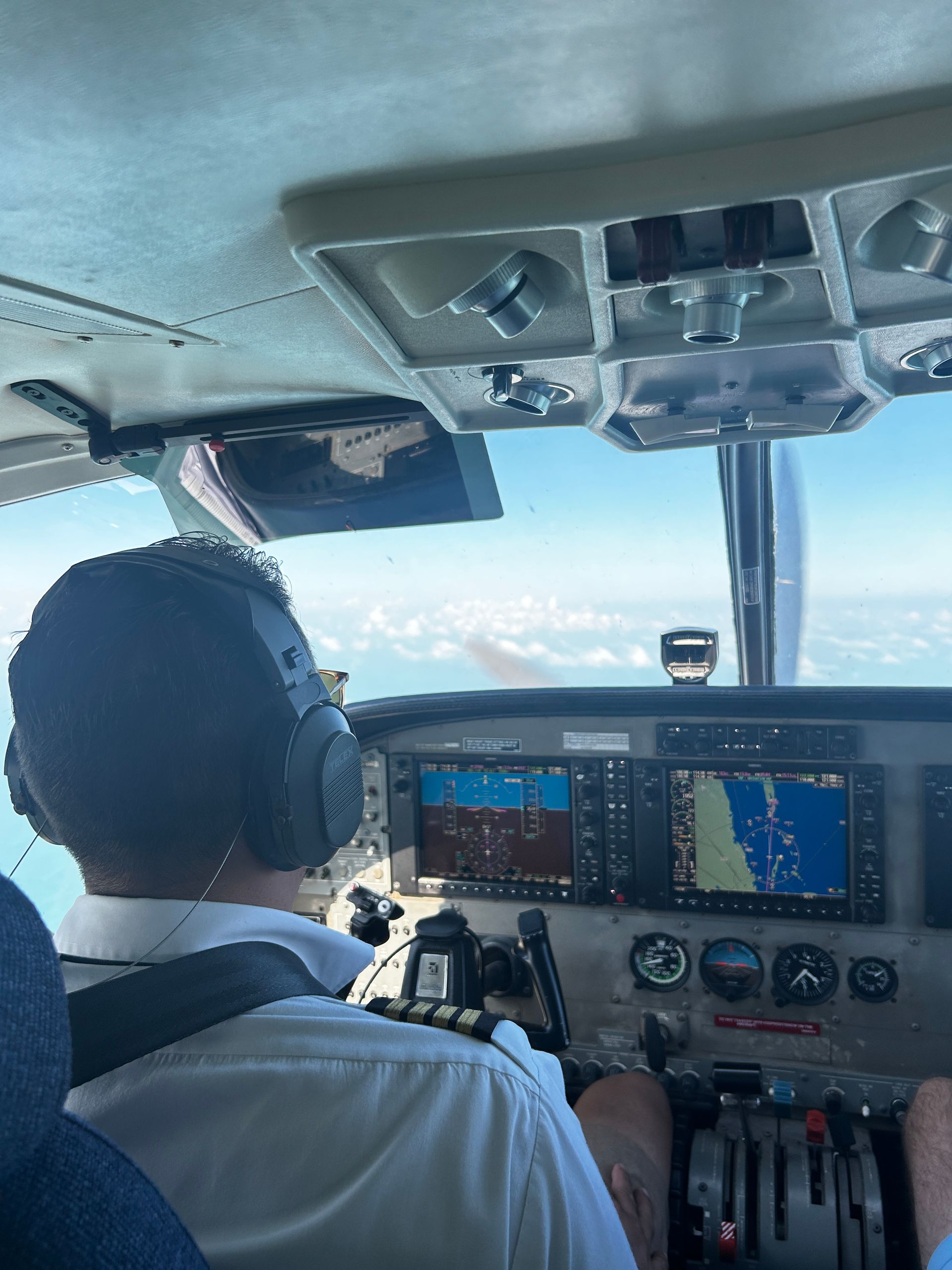 Pilot in cockpit wearing a headset, flying the plane over blue water and sky.