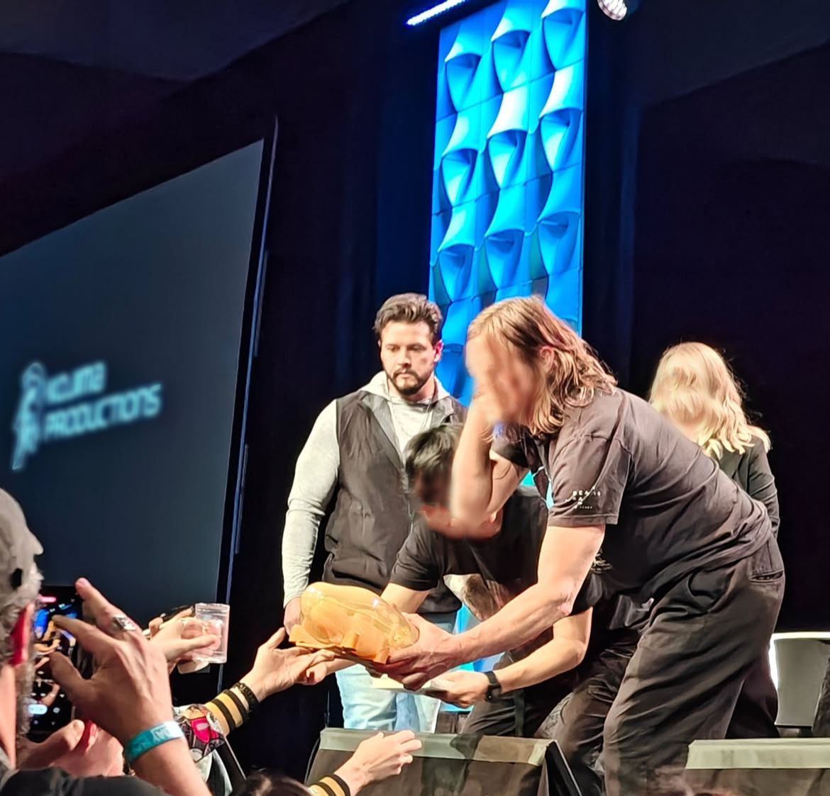People on stage, one holding large bread, handing it to someone as a crowd looks on. Blue and white backdrop.