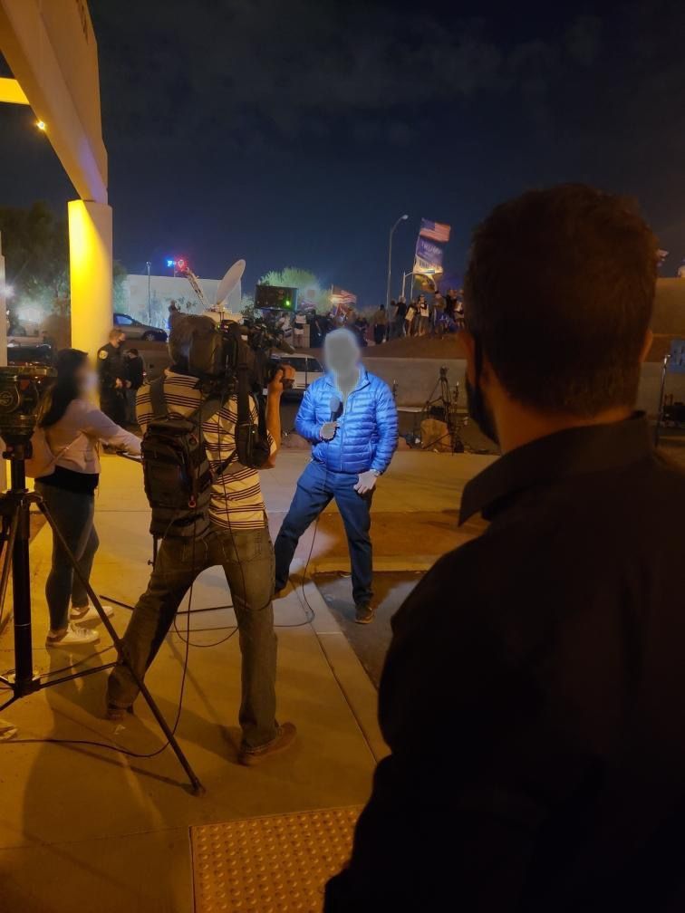 News crew filming reporter outside at night. Reporter in blue jacket speaks to camera.