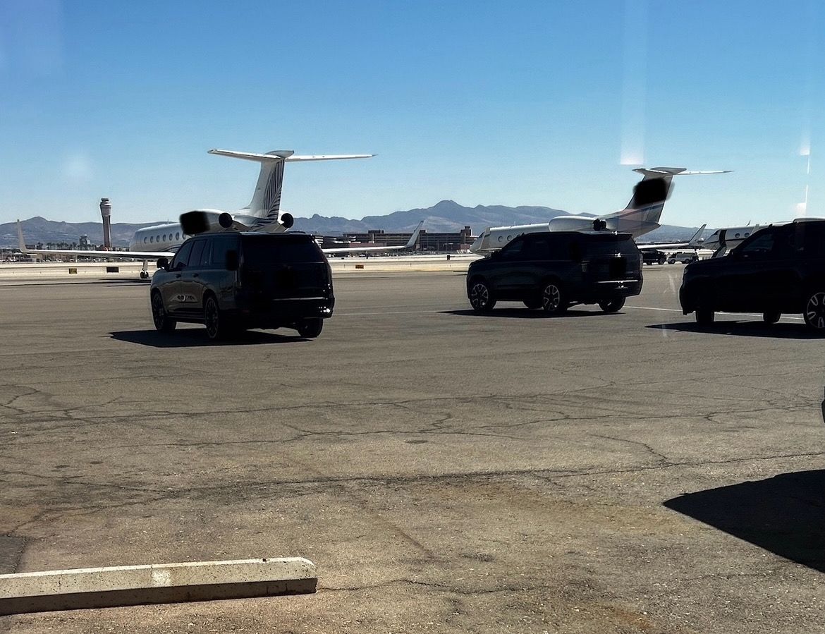 Black SUVs on an airport tarmac with a plane and mountains in the background. Blue sky.