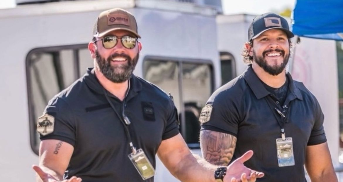 Two men in black shirts with hats, smiling, outdoors.