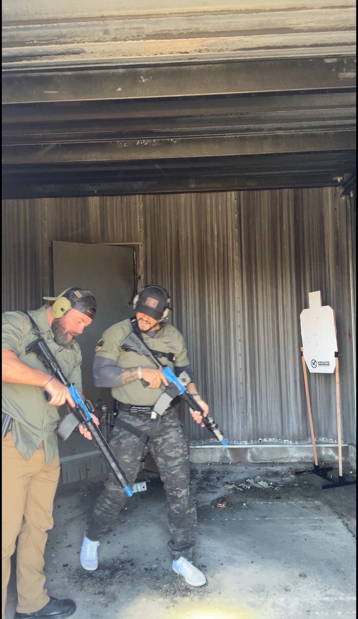 Two men practice shooting rifles at an outdoor range. One wears camo pants, the other khaki. Both have rifles and ear protection.