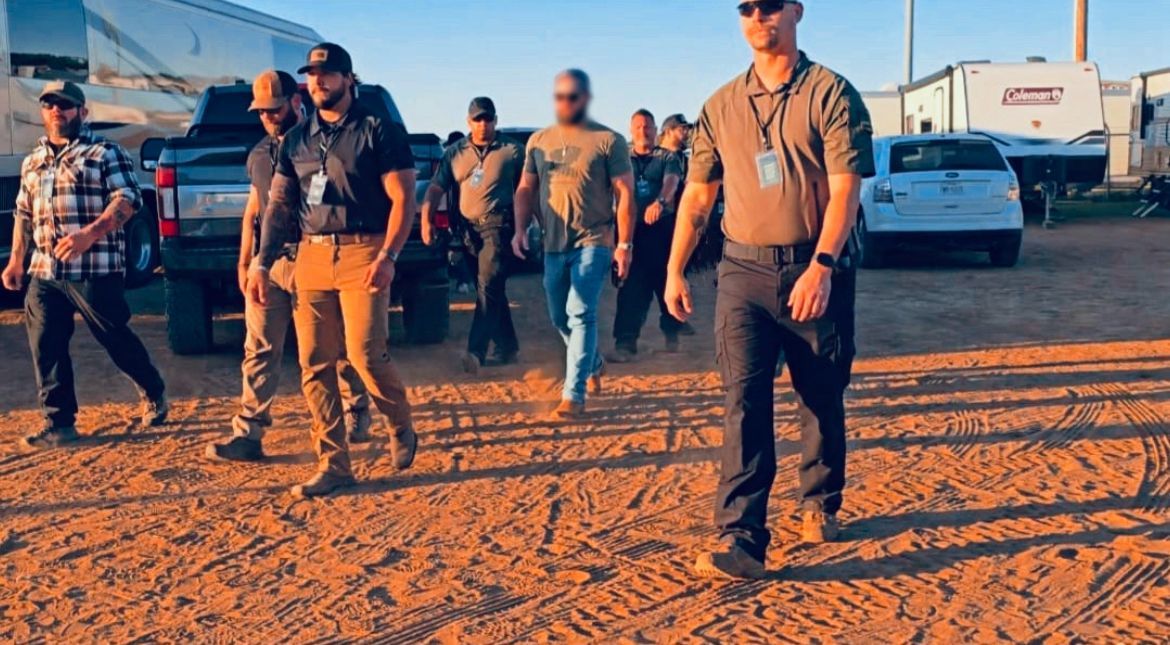 Group of men walking in a dirt lot near trailers and vehicles, wearing hats and sunglasses on a sunny day.