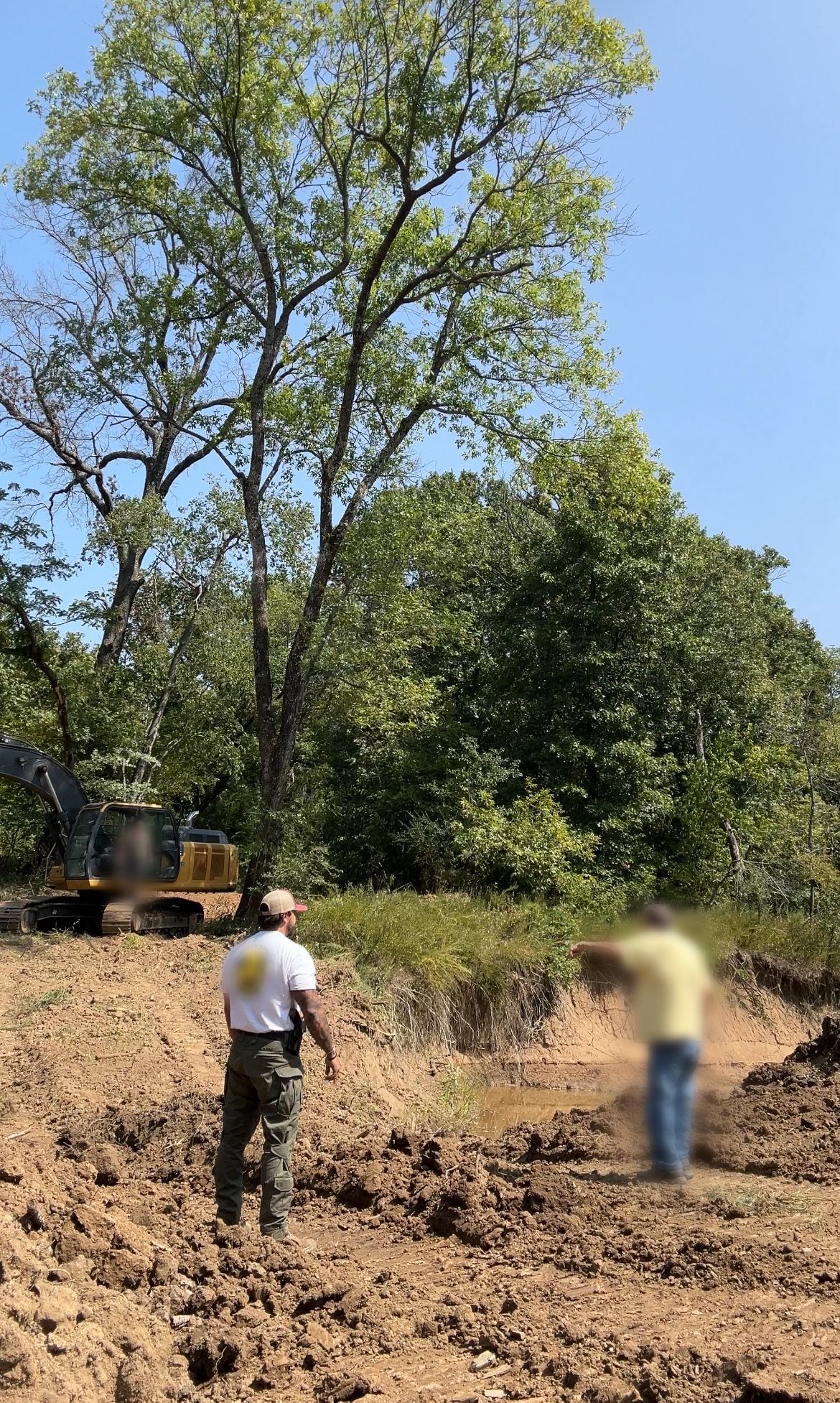 Man in uniform and another blurred person survey a construction site with trees and an excavator.
