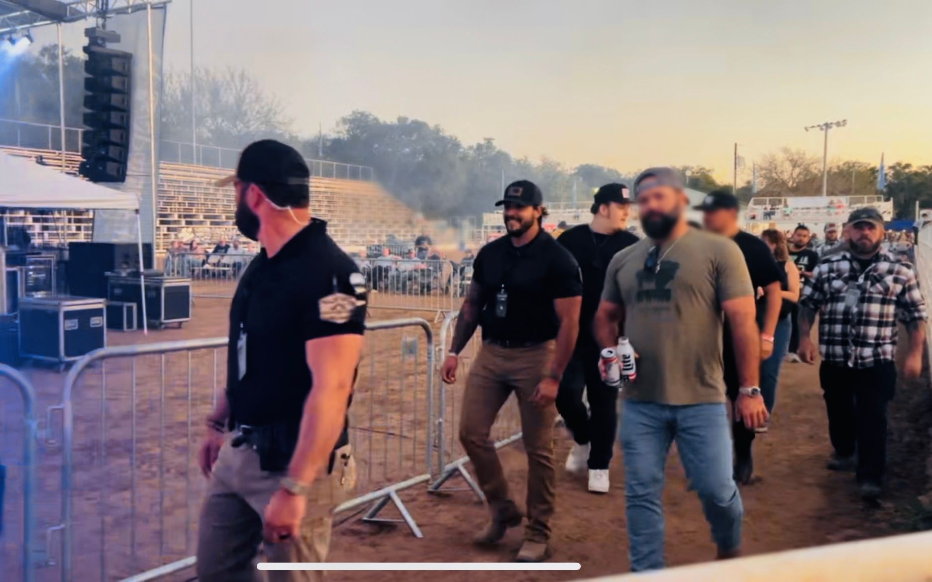Men walking past stage setup at outdoor event; some with tattoos, wearing black shirts, jeans, and sunglasses.
