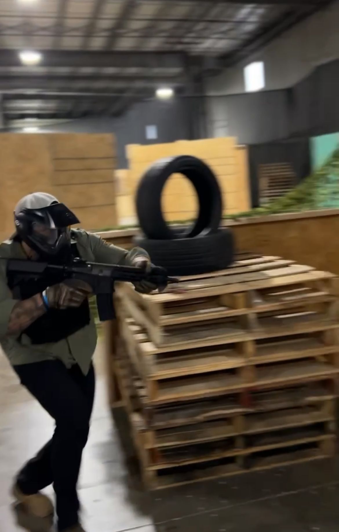 Person in tactical gear aims a rifle, running past pallets and tires inside a warehouse.