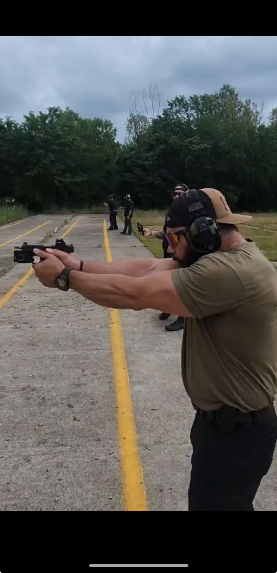 Man aiming a handgun at a shooting range, wearing ear protection and a cap. Other people visible in the background.