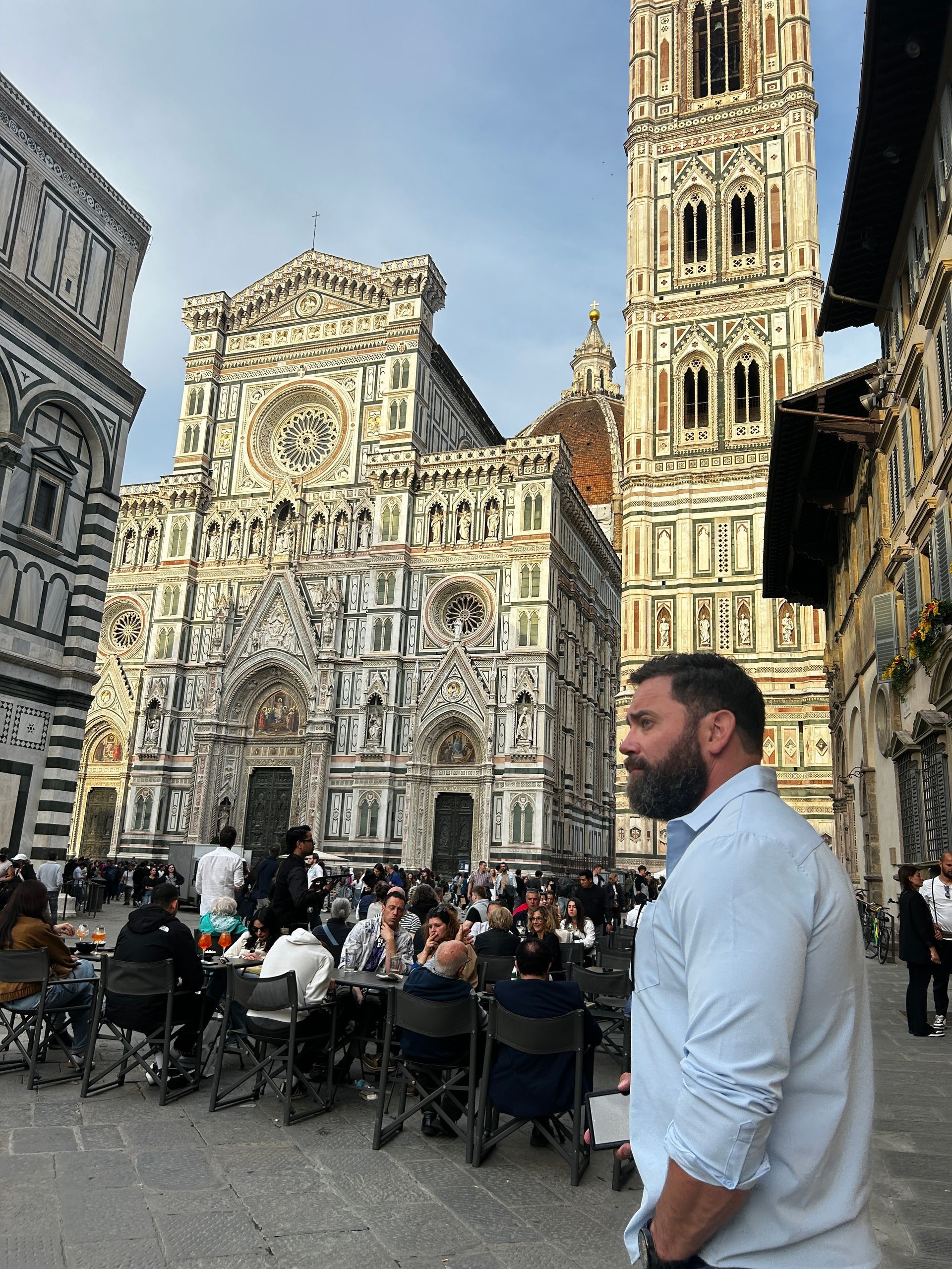 Man in light blue shirt stands before Florence Cathedral, people seated at outdoor tables.