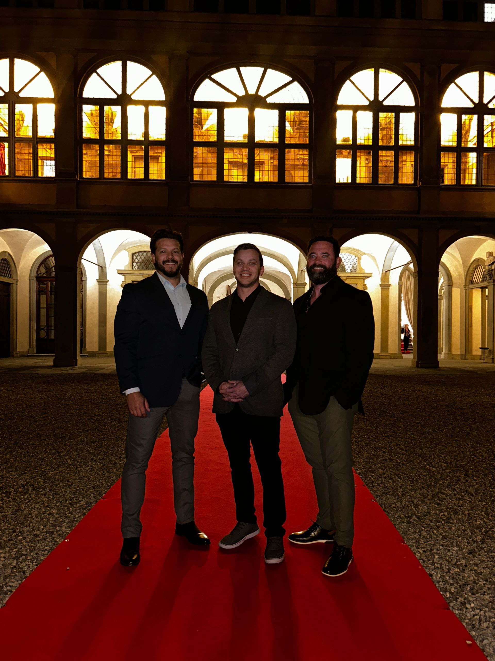 Three men stand on a red carpet in front of an arched building at night.