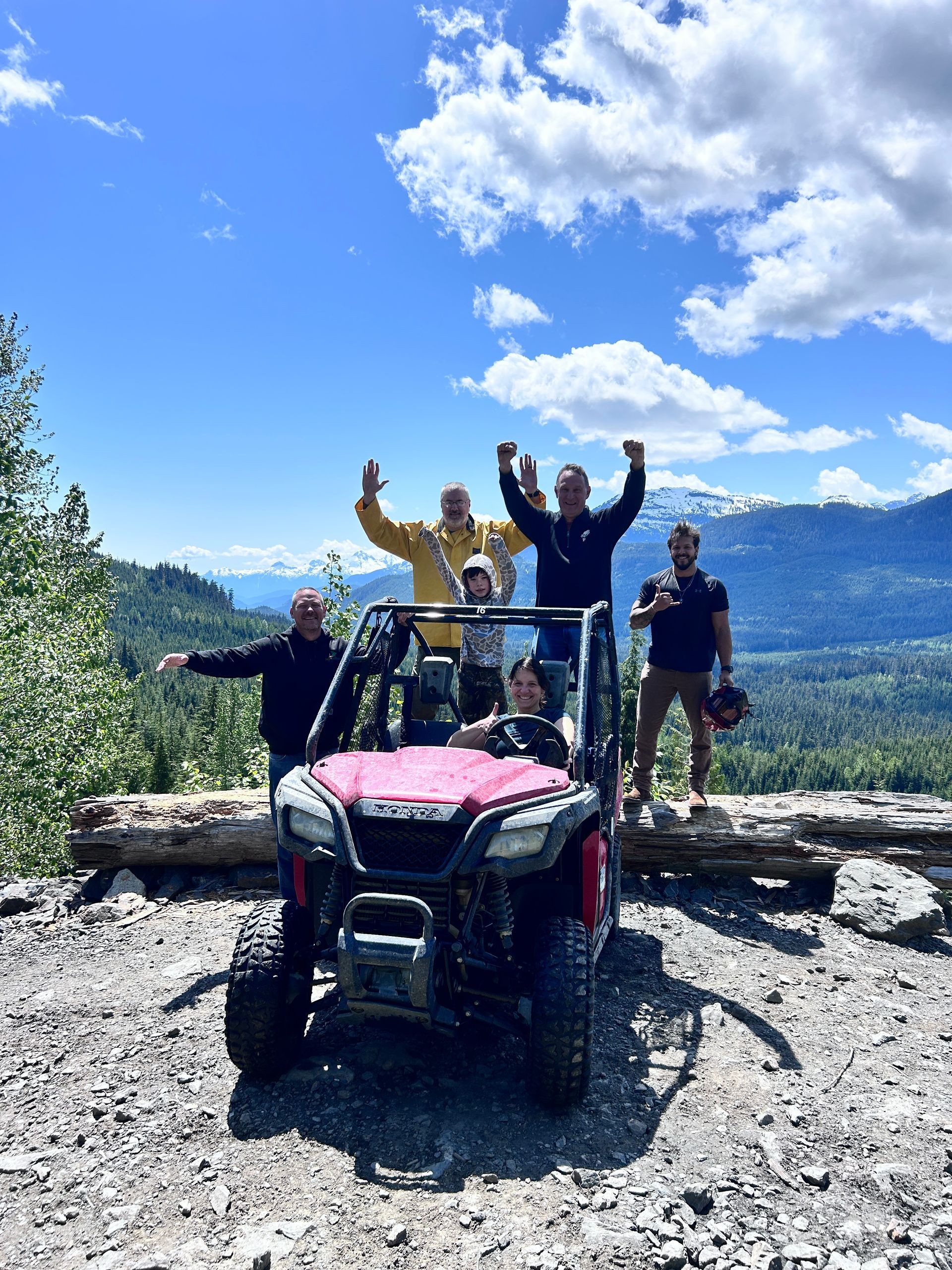 Group of people celebrating in an off-road vehicle, posing against a mountain backdrop on a sunny day.