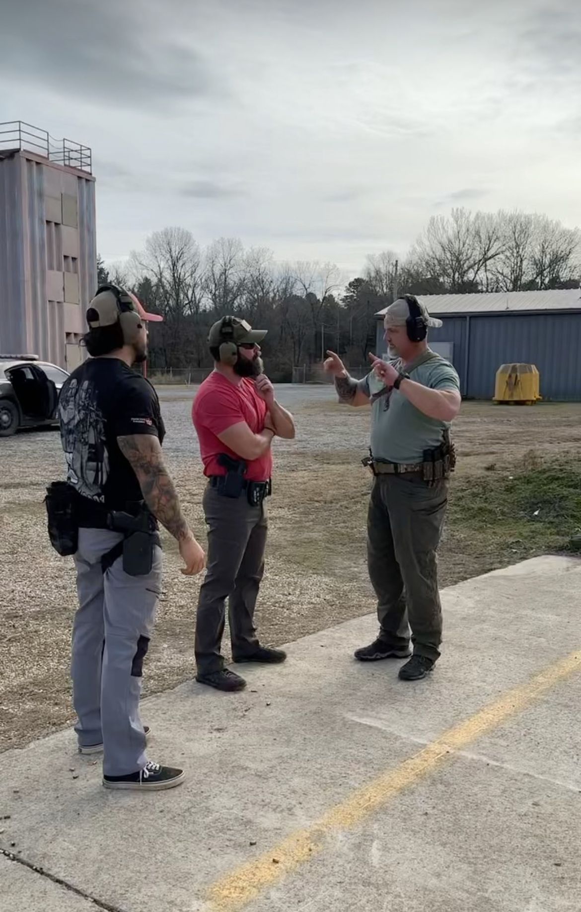 Three men in tactical gear on a shooting range, one gesturing as he speaks, likely instructing.