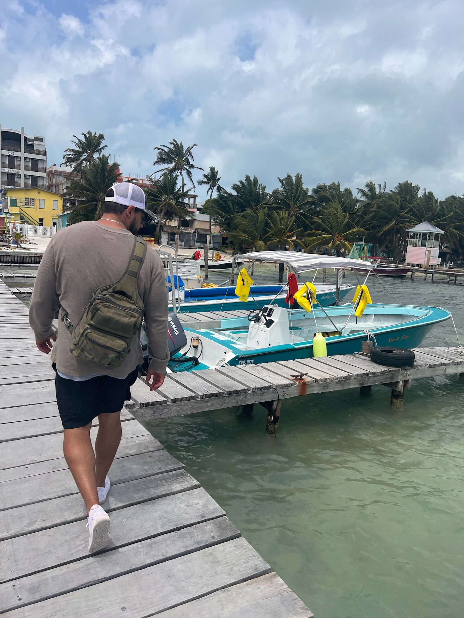 Man walking on a wooden dock toward turquoise boats. Green water, buildings, and trees in the background.