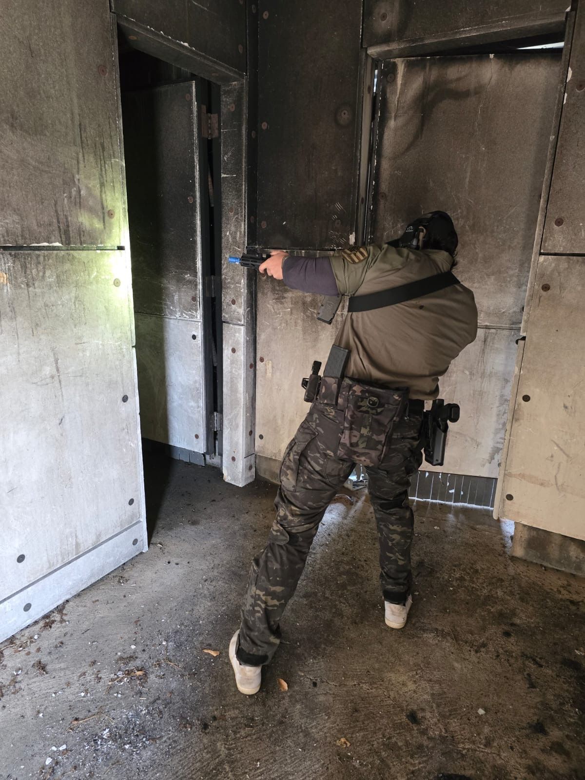 Person in tactical gear aiming a handgun indoors with burned walls and doorway.