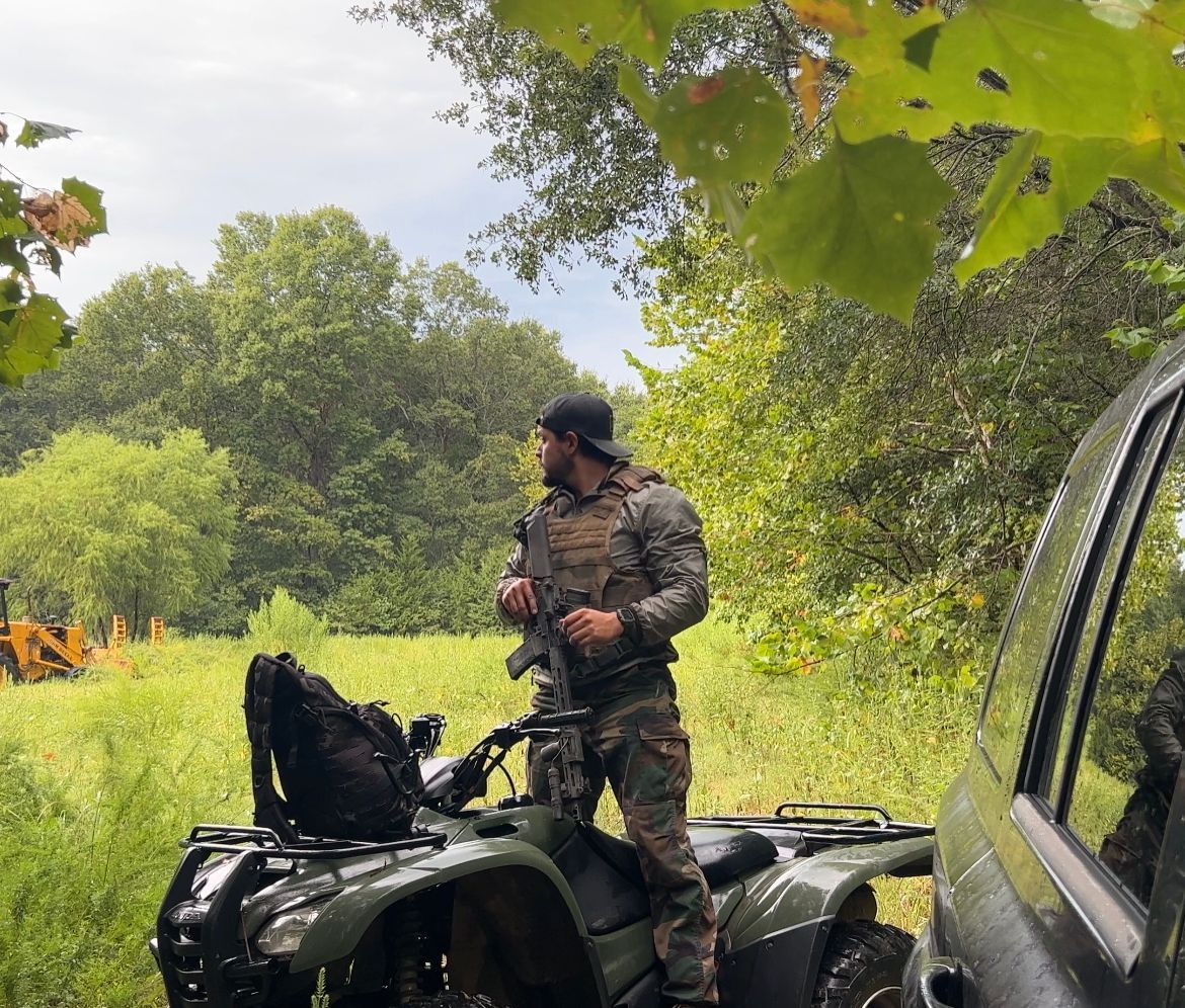Man in camo gear stands on ATV, looking out at a field, near a vehicle and trees.