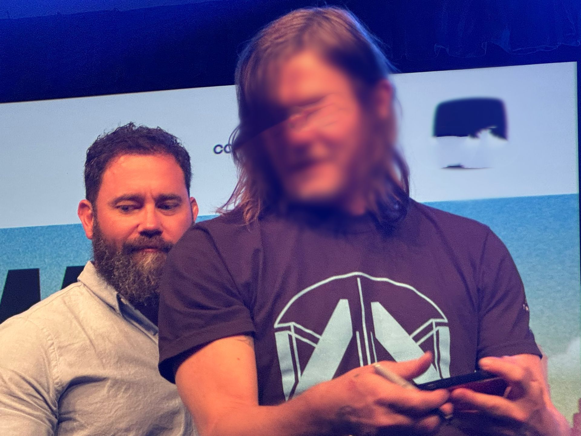 Man with beard and blurry face, holding phone, signing something while another man looks on, indoors.