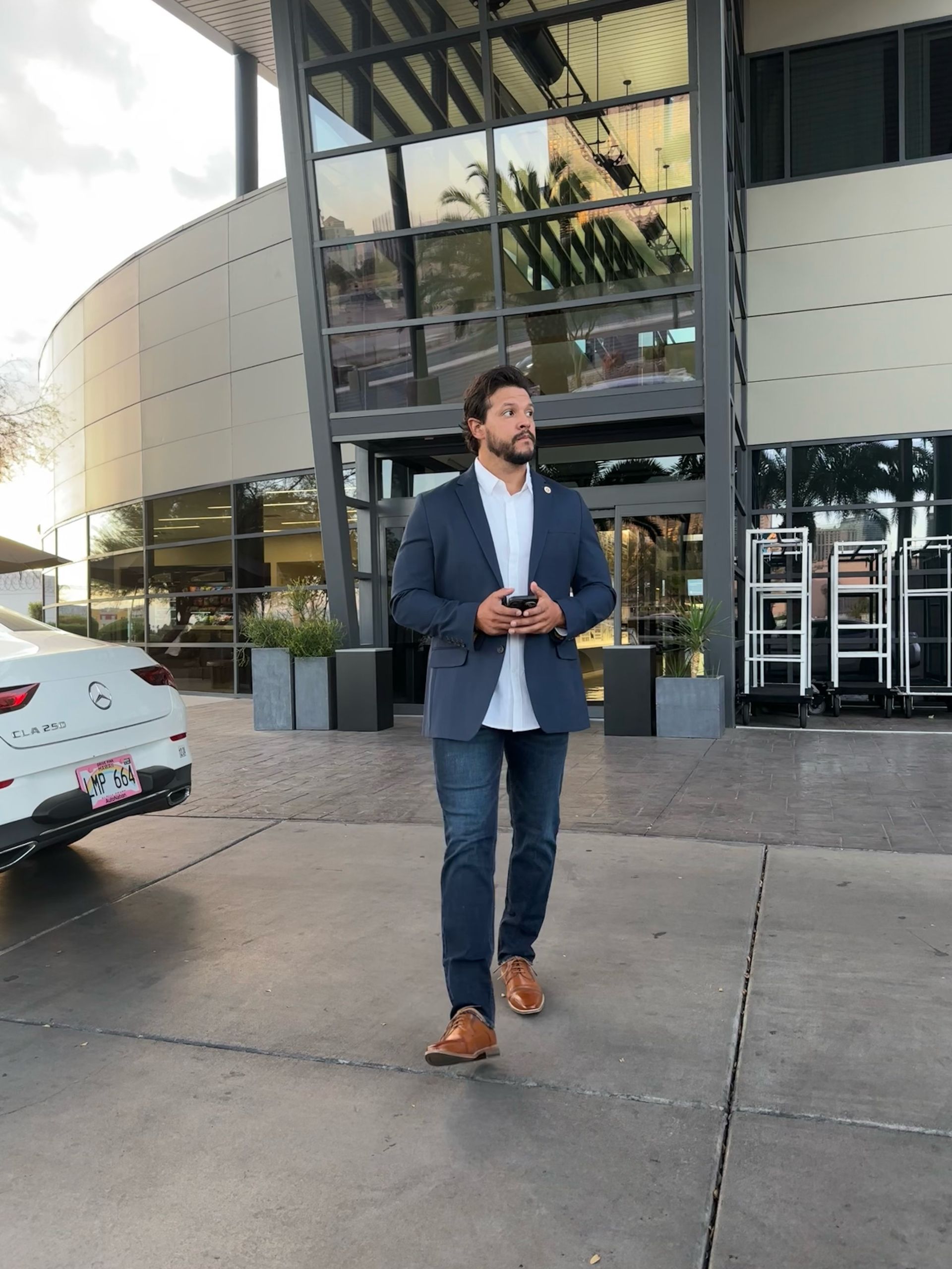 Man in blue blazer and jeans walks toward building entrance, holding phone.
