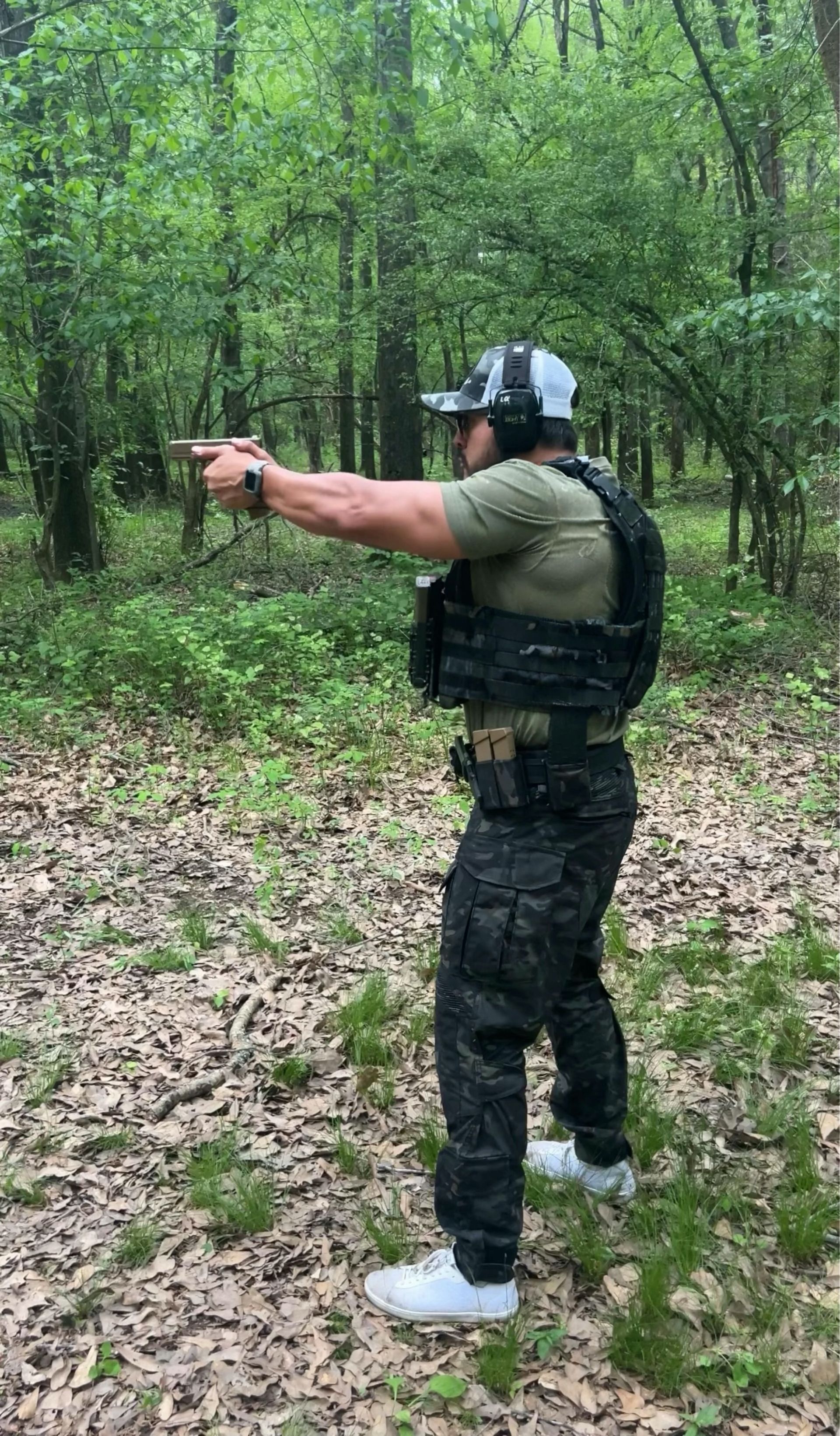 Man in tactical gear aiming a handgun in a wooded area, ready to shoot.