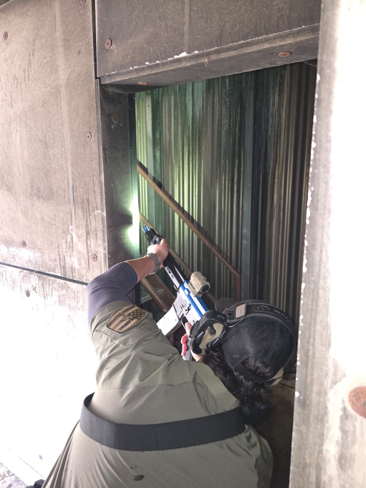 Person holding rifle, aiming into a metal enclosure. They wear a uniform, face mask and a flashlight.