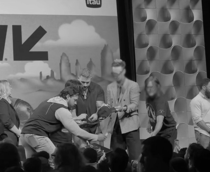 People on stage, one appears to be handing a hat to another. Large arrow backdrop, crowd below.