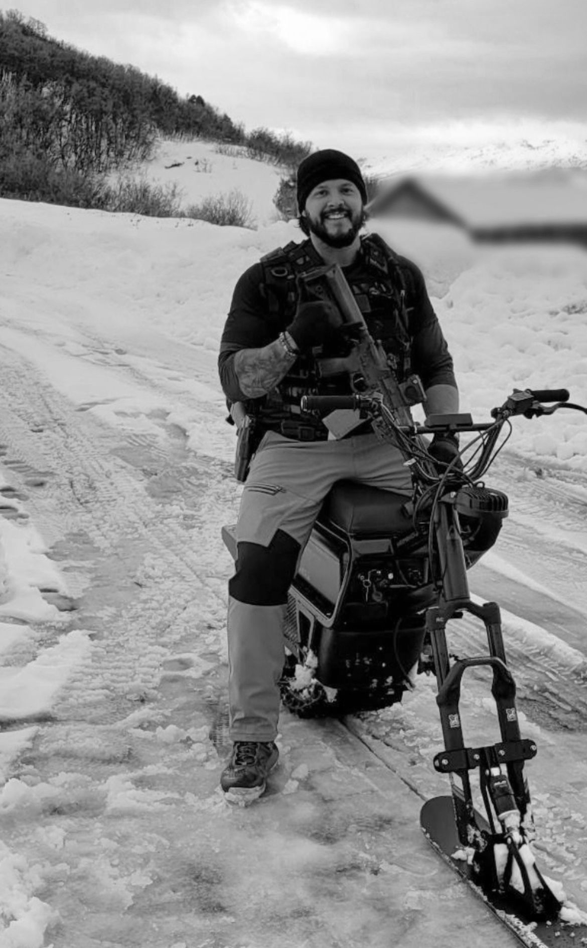 Man on snow bike, rifle in hand, smiles at camera in snowy field.