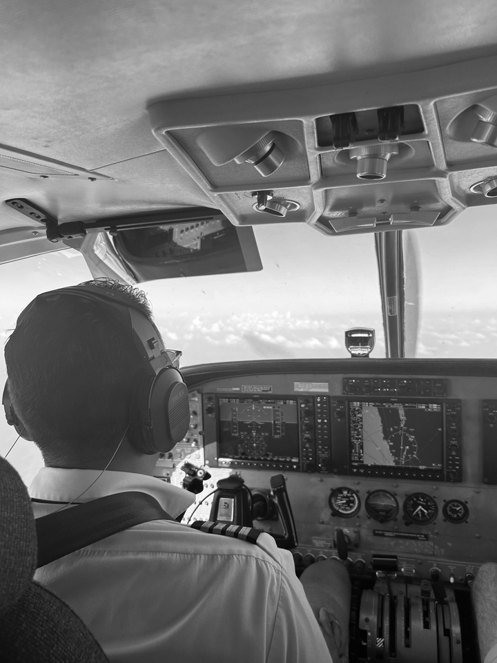Pilot in cockpit, wearing headset, looking at flight instruments. Sunny sky visible through window.