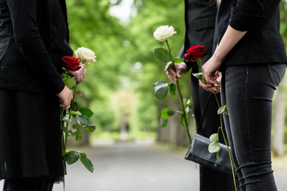 Persone vestite di nero tengono in mano rose rosse e bianche, in piedi lungo un sentiero in un cimitero.