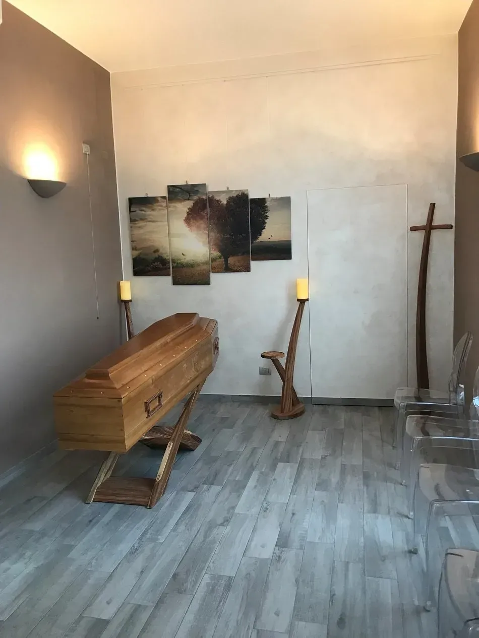 Una bara su un supporto in un'agenzia di pompe funebri, con candele, una croce di legno e opere d'arte.